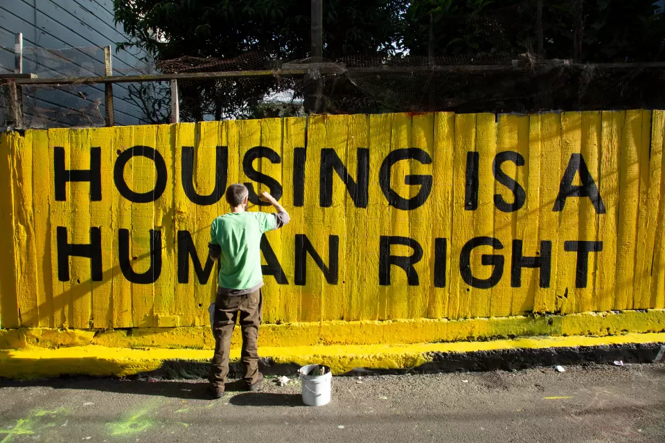 Man painting "HOUSING IS A HUMAN RIGHT" mural in black paint on bright yellow fence